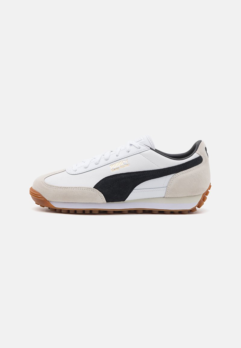 Zapatilla blanca con una franja de ante negro, detalles de ante gris y una suela de goma con un patrón texturizado. Logotipo dorado en el lateral.
