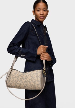 Mujer con chaqueta y falda de mezclilla oscura, sosteniendo un bolso beige con estampado y dos asas, sobre un fondo claro liso.