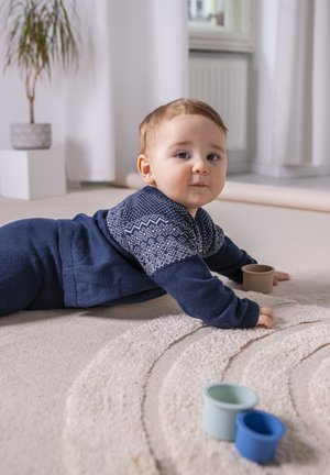 Een baby in een marineblauwe trui met een patroon kruipt op een gestructureerd, licht tapijt. Drie kleurrijke, ronde plastic bekers liggen in de buurt.