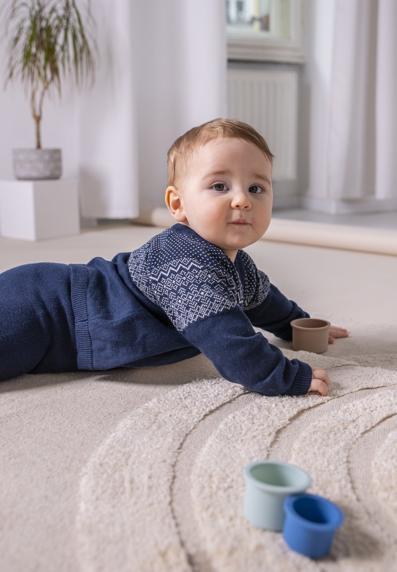 Ein Baby in einem marineblauen Pullover mit einem Muster krabbelt auf einem strukturierten, hellen Teppich. Drei farbige, runde Plastikbecher sind in der Nähe.