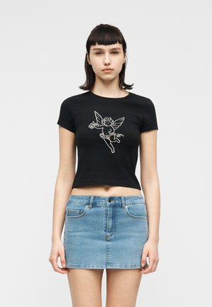 Junge Frau mit kurzen dunklen Haaren, die ein schwarzes, kurz geschnittenes T-Shirt mit einer weißen Cherub-Grafik und einen hellblauen Denim-Minirock trägt.