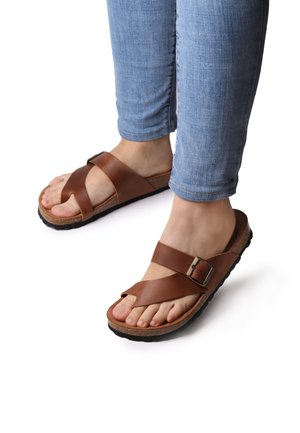 Brown leather sandals with cross straps, adjustable buckle, cork footbed, and textured black rubber sole, paired with fitted light blue jeans.