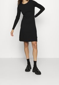 Robe noire à manches longues avec un col rond, une coupe ajustée et des fentes sur les côtés. Associée à des mocassins noirs épais et des chaussettes hauteur cheville.