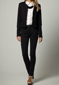 Černá zkrácená bunda přes bílou košili, v kombinaci s černými skinny džíny a hnědými šněrovacími botami. Bunda má zapínání na knoflíky.