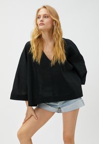 Schwarzes, lässig geschnittenes Leinenoberteil mit V-Ausschnitt und weiten Ärmeln. Kombiniert mit leichten Denim-Shorts, die einen entspannten, lässigen Look zeigen.