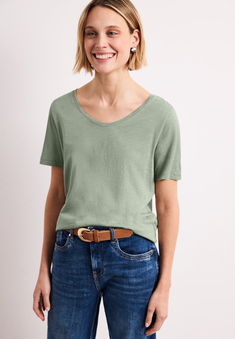 Femme souriante aux cheveux blonds courts portant un t-shirt vert clair à manches courtes, un jean bleu et une ceinture marron, sur fond uni.