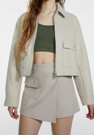 Woman wearing a beige cropped jacket over a green ribbed crop top and beige asymmetrical skirt with a large front pocket.
