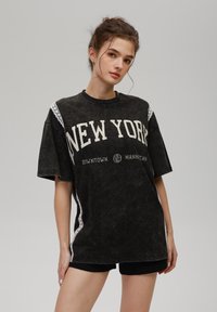 Ung kvinne med håret samlet bak, iført en oversized svart New York T-skjorte og svarte shortser foran en ensfarget grå bakgrunn.