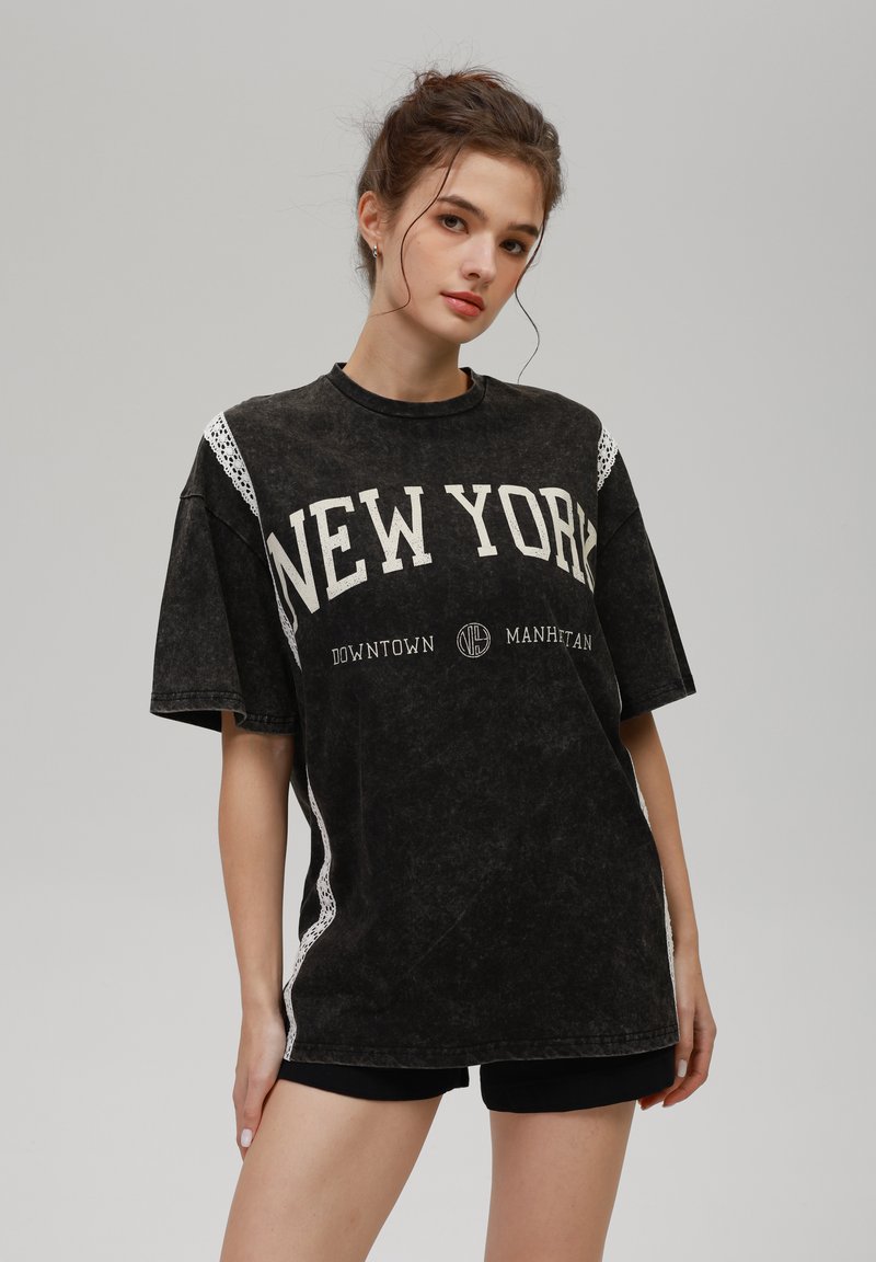 Ung kvinne med håret samlet bak, iført en oversized svart New York T-skjorte og svarte shortser foran en ensfarget grå bakgrunn.