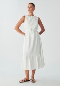 White sleeveless dress with a gathered waist and ruffled hem, made of soft fabric. Model wears black strappy sandals.