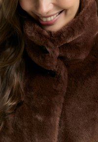 Brauner Kunstpelzmantel mit hohem Kragen, zeichnet sich durch eine glatte Textur und Knopfdetails aus. Nahaufnahme des Stoffes und Kragenbereichs.