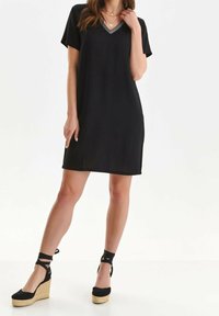 Černé šaty s krátkým rukávem, ve výstřihu do V a geometrickým vzorem na dekoltu; zkombinované s černými espadrilovými klínovými botami. Hladká textura látky.