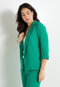 Blazer verde texturizado con solapa muesca, mangas tres cuartos y puños enrollados. Llevado sobre una blusa blanca con un drapeado sutil.