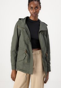 Veste zippée vert olive avec capuche, taille à cordon et accents en métal doré ; fabriquée en tissu durable et doux. Poches avant incluses.