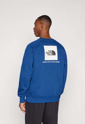 Sudadera azul hecha de tela suave, con un logo cuadrado blanco que incluye el texto "The North Face" y "Never Stop Exploring" en la parte posterior.