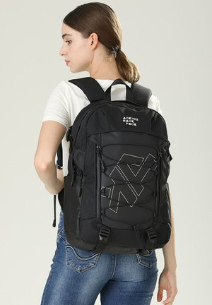 Sac à dos noir avec une finition texturée, présentant un motif géométrique, plusieurs sangles et des cordes bungee à l'avant pour un espace de rangement supplémentaire.