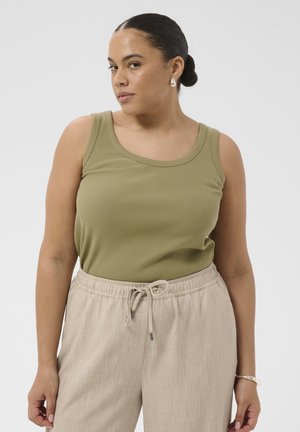 Femeie cu păr închis la culoare prins în coc jos poartă un top verde măsliniu fără mâneci, pantaloni bej cu șnur, cercei de perlă și o brățară argintie, stând neutră.