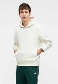 Jovem com um hoodie creme, com as mãos nos bolsos da frente e calças de treino verdes, em pé contra um fundo branco liso.