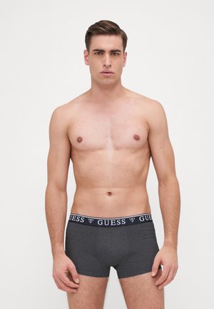 Szürke boxeralsó sötét derékrésszel, amelyen fehér színű "GUESS" logó található. Az anyag sima és testhez simuló hatású. A modell előrefelé néz.