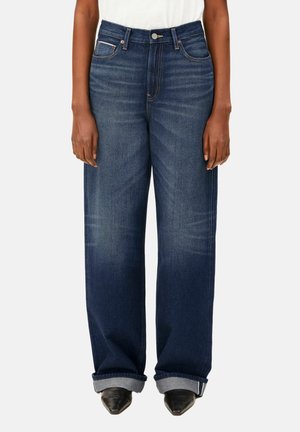 Person, die hoch taillierte, dunkelblaue Jeans mit weitem Bein und Umschlägen trägt, kombiniert mit schwarzen spitzen Schuhen und einem weißen Oberteil, stehend.