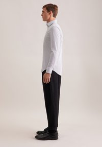 Camicia bianca a maniche lunghe con un sottile motivo a pois, abbinata a pantaloni neri e scarpe eleganti nere, vista di lato.