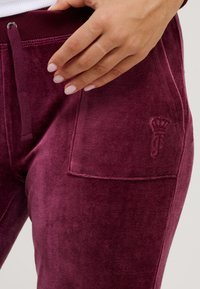 Burgundy velour joggers featuring a side pocket, elastic waistband with drawstring, and embossed logo detail near the pocket.