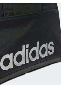 Musta Adidas kott, millel on matte tekstuur, suur valge logo ja struktureeritud kuju koos läikiva alumise aktsendiga.