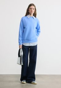 Μια γυναίκα στέκεται φορώντας ανοιχτό μπλε φούτερ πάνω από ριγέ πουκάμισο, φαρδιά σκούρα τζιν, ασημί αθλητικά παπούτσια και κρατώντας μια μεταλλική ασημένια τσάντα.