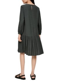 Dunkelgrünes Kleid mit langen Puffärmel, einem eingekräuselten Rock, subtil gemusterter Textur und einem Schlüsselspaltverschluss auf der Rückseite. Getragen mit schwarzen Sandalen.