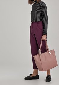 Borsa a spalla rosa con due manici, forma rettangolare, realizzata in materiale liscio. Abbinata a un top a righe bianche e nere e pantaloni viola. Scarpe nere.