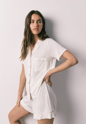 Vrouw met lang bruin haar, gekleed in een witte kortarmige blouse met knopen en bijpassende shorts, staand met de hand op de heup tegen een effen muur.