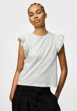 Donna con capelli intrecciati che indossa una canotta bianca con bordo in pizzo arricciato e pantaloni neri, in piedi con le mani nelle tasche.