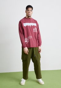 Langermet burgunder skjorte med hvite grafiske trykk og grønne cargobukser; har snøring i livet og lommer. Hvite sneakers fullfører looken.