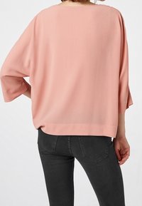 Blusa suelta de color rosa suave con mangas de tres cuartos, cuello redondo y textura suave, combinada con jeans ajustados oscuros.