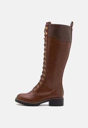 Botte en cuir marron haute jusqu'au genou avec des œillets dorés, un devant lacé, un talon robuste et une finition texturée. Elle présente un revers marron contrastant en haut.