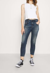 Top corto blanco de tirantes con dobladillo ondulado y botones dorados, combinado con jeans ajustados azul oscuro que tienen bolsillos frontales y un acabado desgastado.