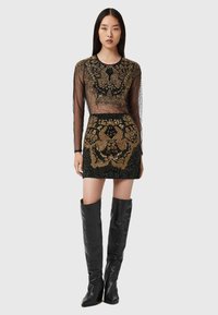 Przezroczysta bluzka z długim rękawem i dopasowana mini spódnica z złotym haftem na czarnym materiale; zestawione z czarnymi skórzanymi kozakami do kolan.