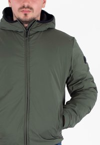 Veste à capuche verte avec une texture lisse, fermeture éclair à l'avant et un patch rond noir sur l'épaule gauche.