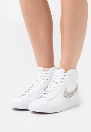 Gambe con indosso scarpe da ginnastica bianche alte con lacci bianchi e logo a forma di swoosh grigio con motivo a zebra su sfondo bianco.