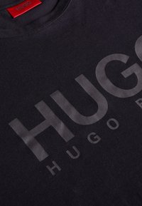 Sort bomulds T-shirt med et stort, glinsende "HUGO" logo i subtil grå. Rund halsudskæring med standard syninger og et rødt mærke på bagsiden.