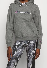 Felpa grigia con tasca a marsupio, cappuccio a strati con cordoncino e logo "Champion" ricamato in bianco e rosso. Indossata con leggings a fantasia.