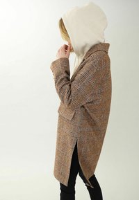 Ruskea ruudullinen takki, jossa on muotoiltu malli, sivuhalkiot ja kaulus, yhdistettynä vaaleanbeigeen huppariin sen alla.