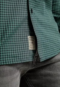 Zielono-czarna koszula w kratę wykonana z tkaniny o teksturowanej powierzchni. Posiada metkę na dole, zestawiona z szarymi dżinsami.