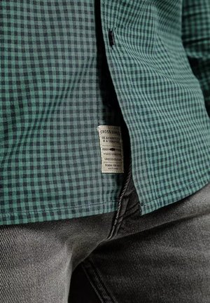 Zielono-czarna koszula w kratę wykonana z tkaniny o teksturowanej powierzchni. Posiada metkę na dole, zestawiona z szarymi dżinsami.