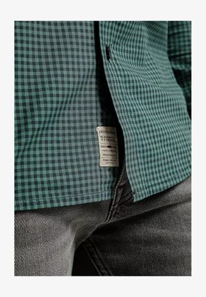 Zielono-czarna koszula w kratę wykonana z tkaniny o teksturowanej powierzchni. Posiada metkę na dole, zestawiona z szarymi dżinsami.