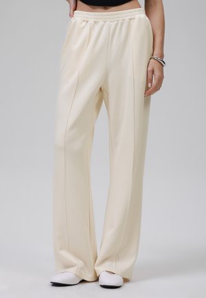 Lichtbeige wijdvallende broek van gladde stof; heeft een elastische tailleband en zijzakken. Gemaakt in combinatie met witte instap schoenen.