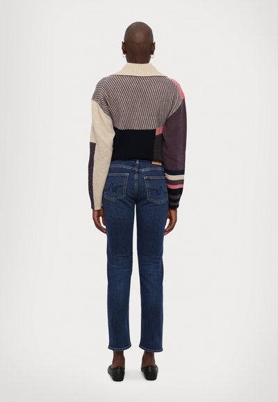 Suéter de punto multicolor con diversos patrones, textura acanalada y cuello alto, combinado con jeans de mezclilla azul oscuro con bolsillos traseros.