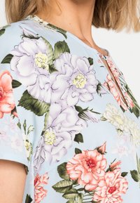Femme portant une chemise bleu clair à manches courtes avec un motif floral en rose, violet, blanc et feuilles vertes.