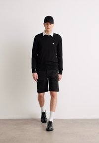 Svart genser over en hvit skjorte med krage, sammen med svarte cargo-shortser og svarte lærsko. Modellen har på seg en svart caps.
