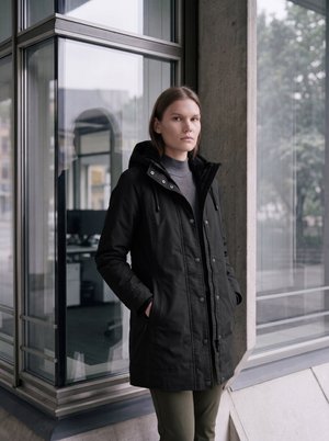 Lühikeste juustega noor inimene seisab toas klaasakna ääres, kandes musta kapuutsiga mantel ja halli kõrge kaelusega kampsunit.